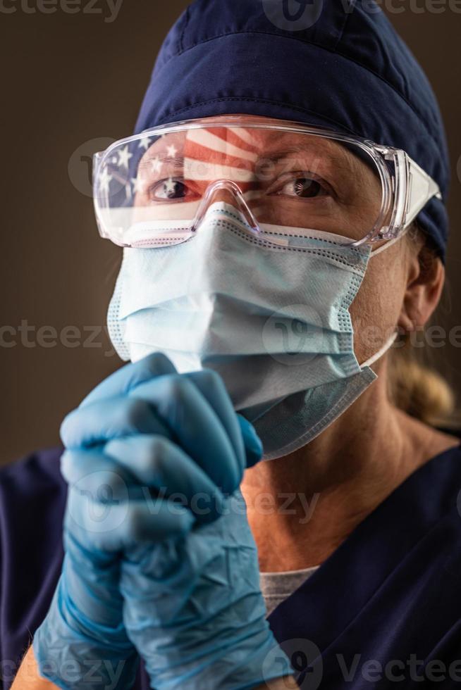 American Flag Reflecting on Distressed Praying Female Medical Worker Wearing Protective Face Mask and Goggles photo