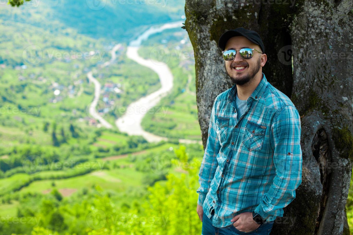 man smiling and posing near tree with mountains landscape from behind photo