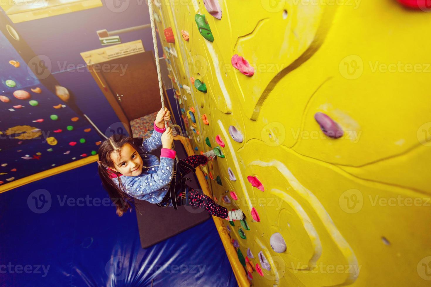 Little baby girl with funny hear style climbing vertical wall and man belaying her from below photo