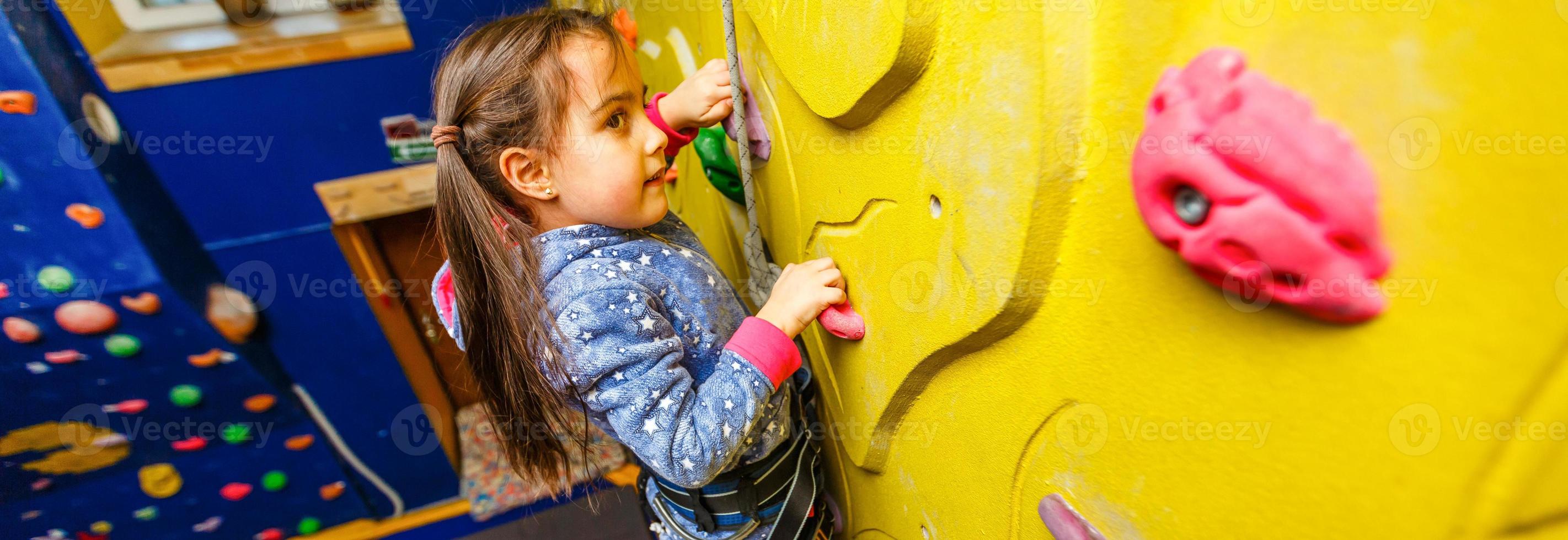 Little baby girl with funny hear style climbing vertical wall and man belaying her from below photo