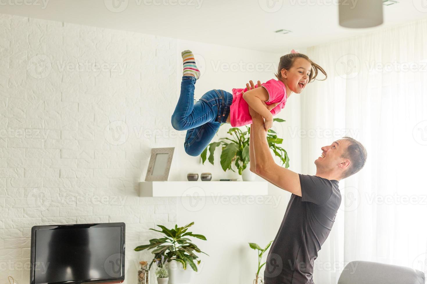 I can fly. Happy dad is lifting up his little daughter while standing in living room. photo