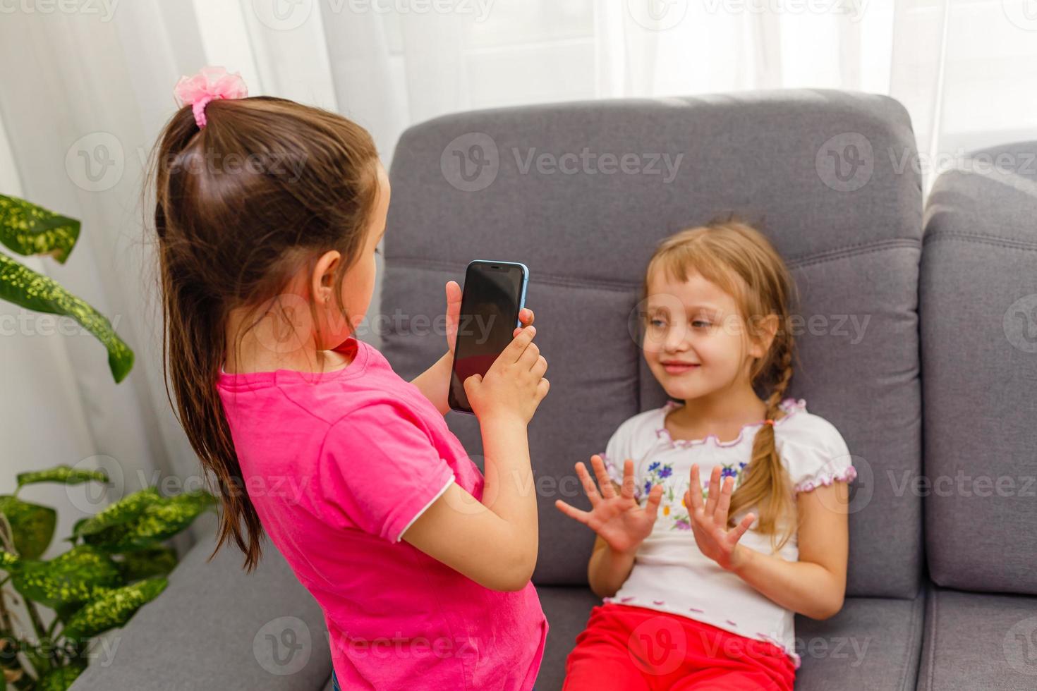 people, children, technology, friends and friendship concept - happy little girls sitting on sofa and taking selfie with smartphone at home. photo