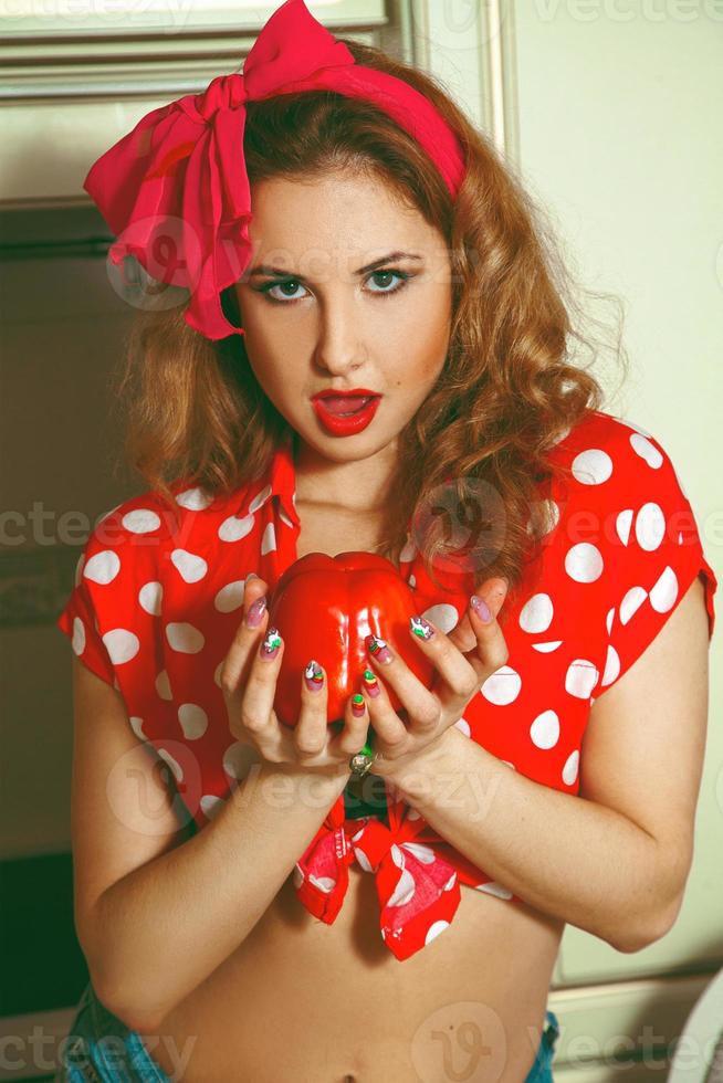 Cute young girl in pinkup style posing in the kitchen with peper in hands photo