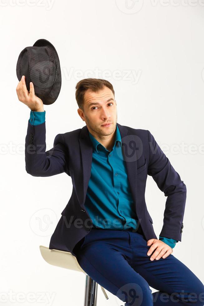 sexy confident man wearing a black tuxedo sitting on a metal chair on light background, photo