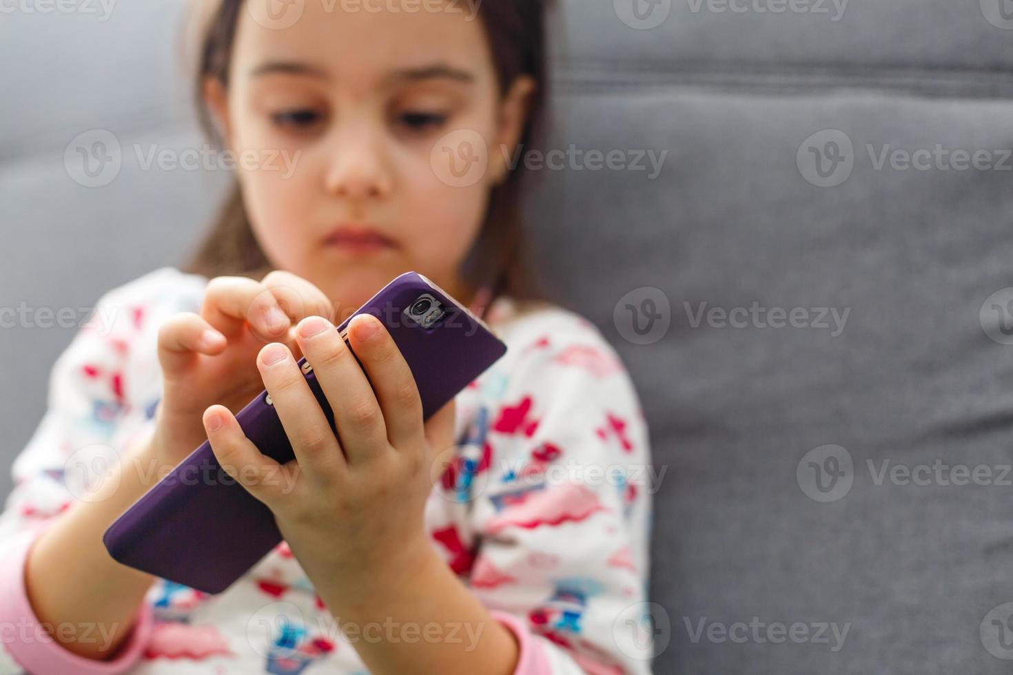 Beautiful little girl playing game or watching video on smartphone mobile. Girl watching cartoons or browsing internet, copy space. Side view portrait of little girl using smartphone while sitting. photo