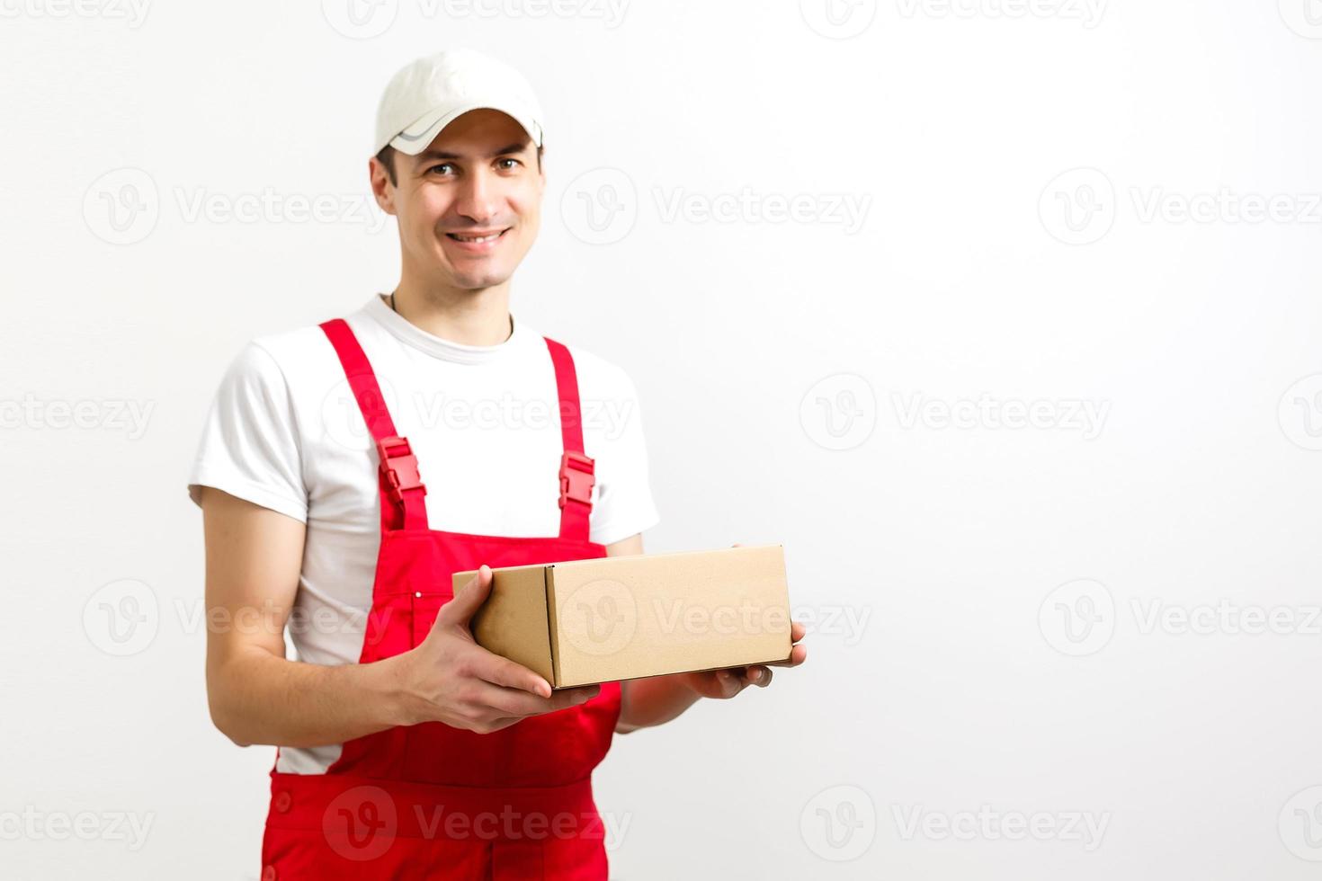 Mago de un joven y feliz repartidor con gorra roja de pie con un buzón de paquetes aislado sobre fondo blanco. foto