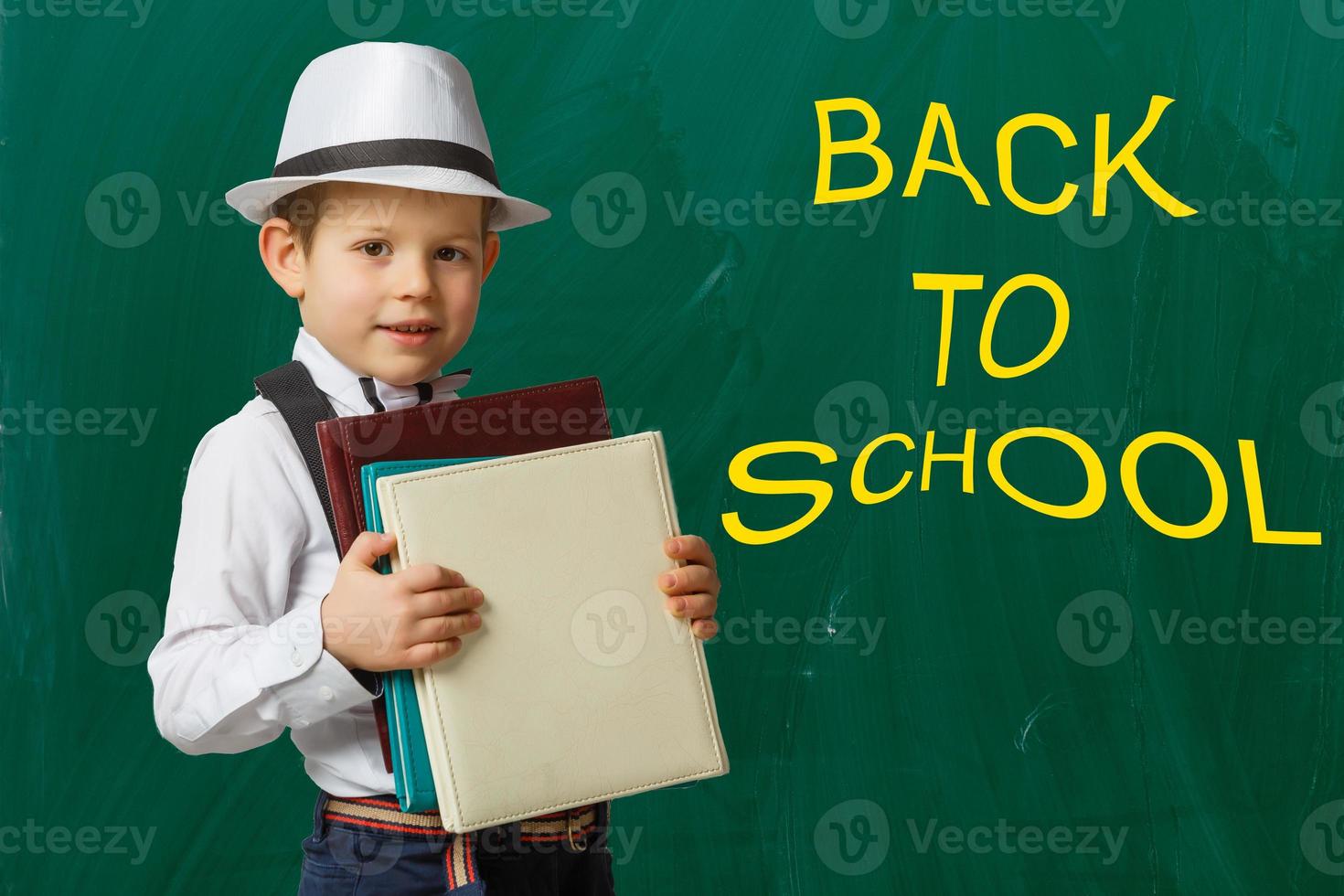 Back to school. Funny little boy in glasses pointing up on blackboard. Child from elementary school with book and bag. Education. photo