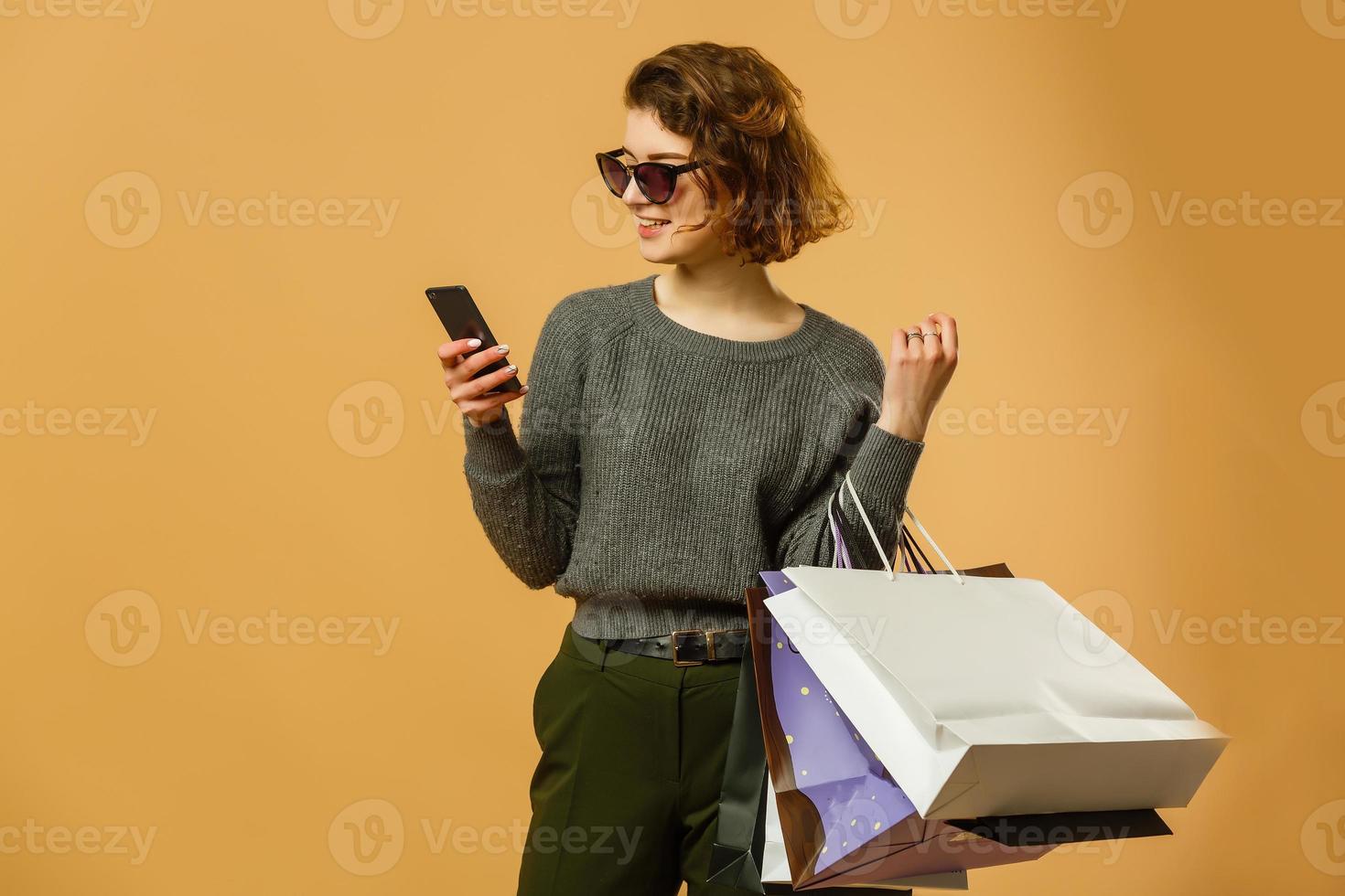 hermosa mujer joven con bolsas de compras usando su teléfono inteligente sobre fondo amarillo. moda de compras adicta a las compras foto