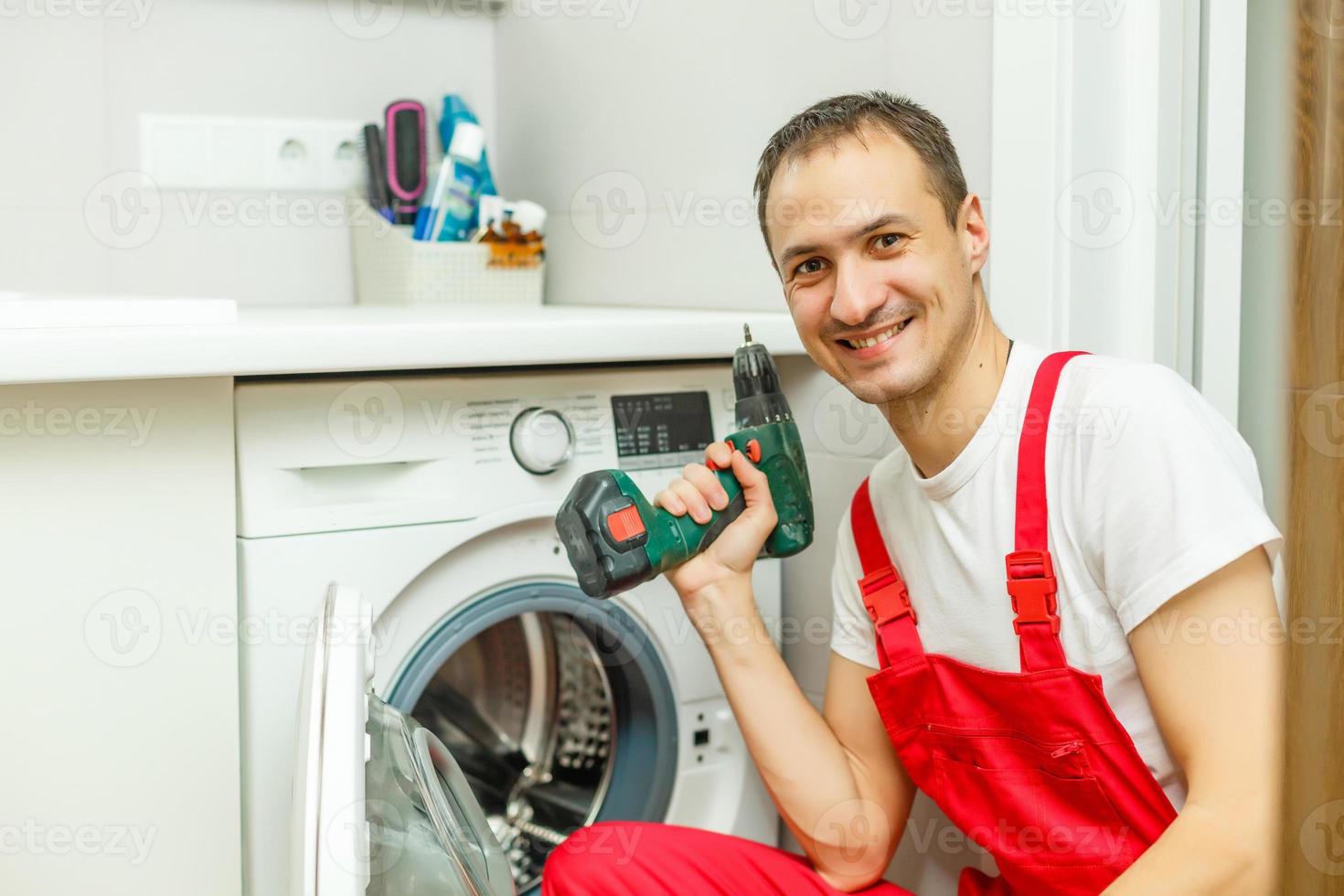 Young attractive smiling worker in uniform fixing washing machine, background photo