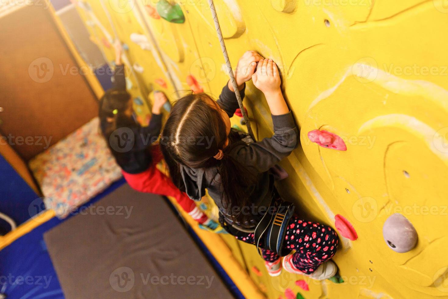 Little baby girl with funny hear style climbing vertical wall and man belaying her from below photo