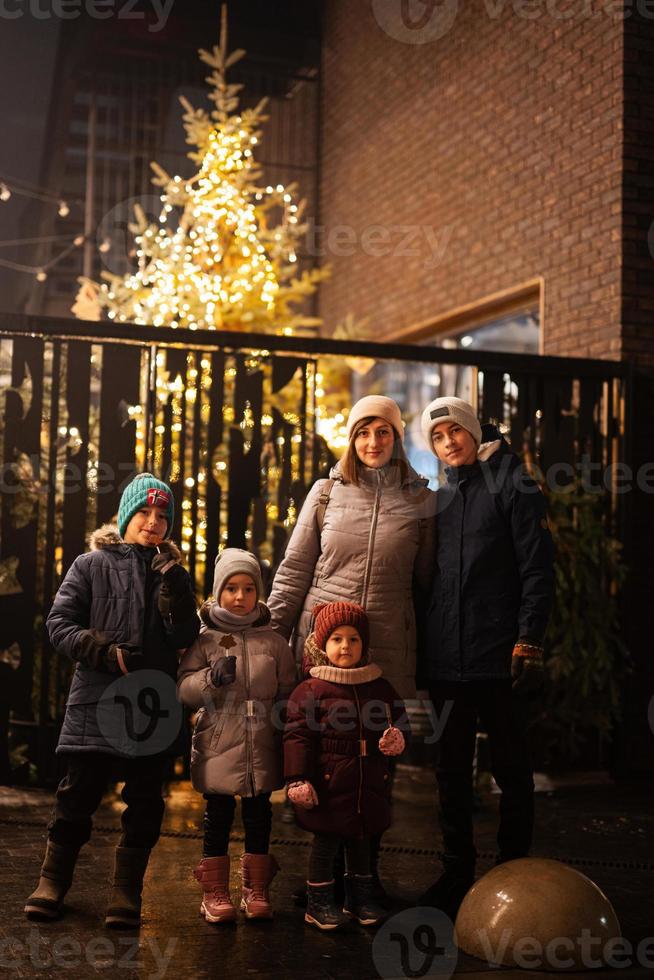 Mother with four kids stand against illuminated Christmas tree outdoor in evening. photo