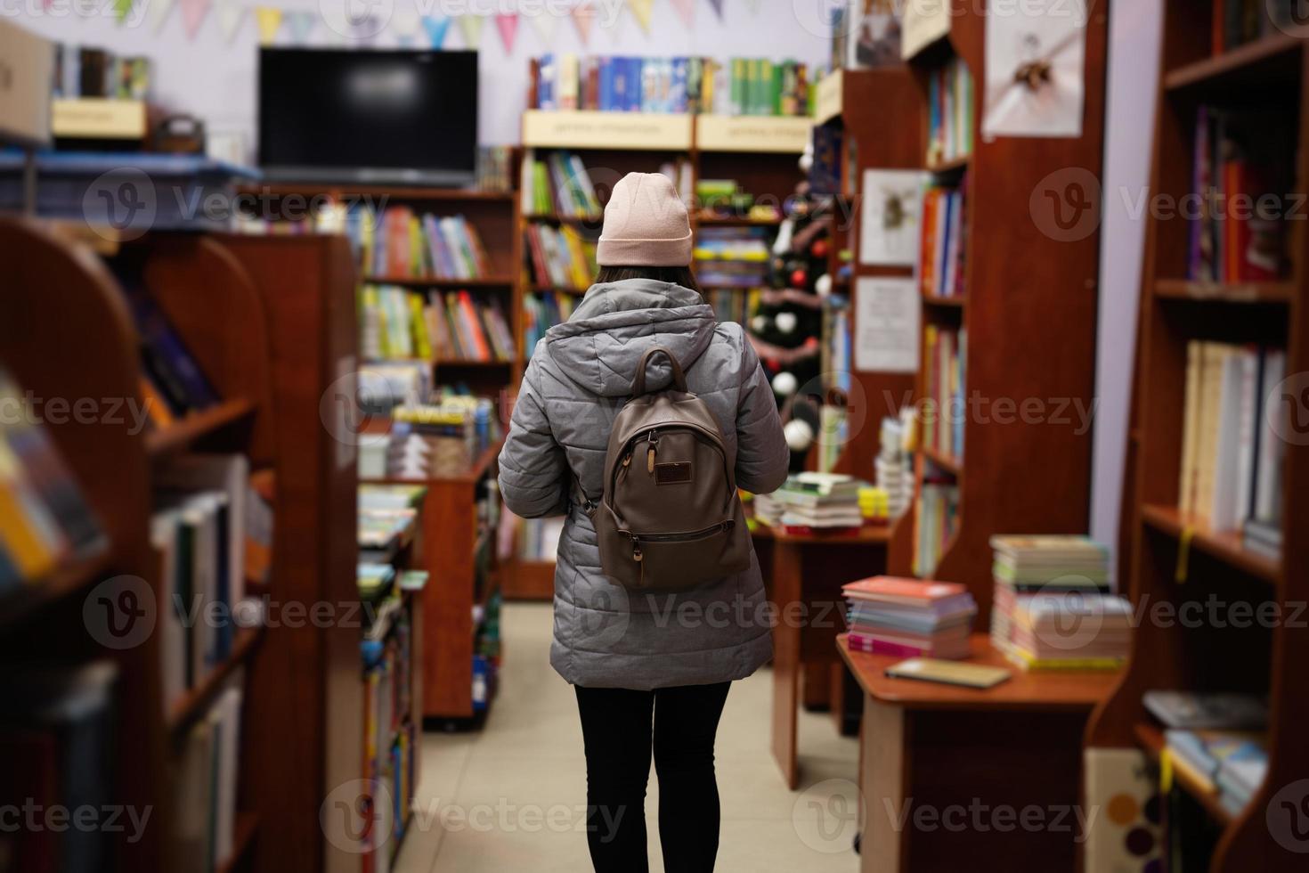 Back of woman in jacket and backpack reaching a book from bookshelf at the library. photo