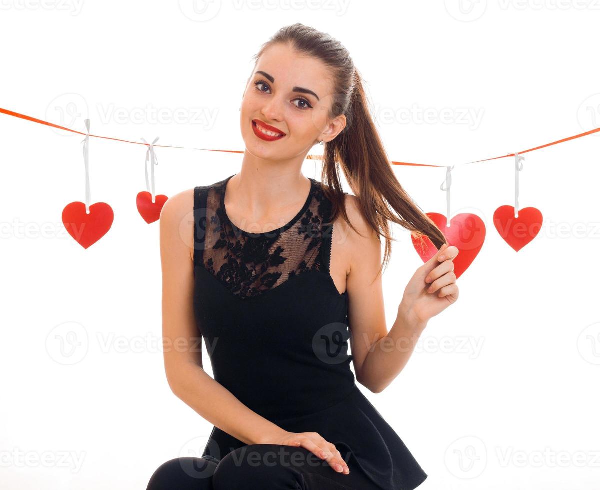 square photo of young cheerful brunette lady with red heart in studio smiling on camera isolated on white background. Valentines Day concept.