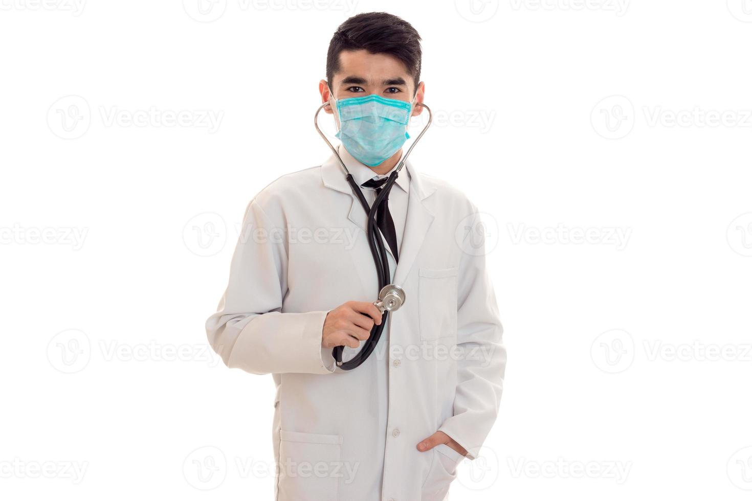 serious brunette male doctor in uniform with stethoscope on his neck and mask posing and looking at the camera isolated on white background photo