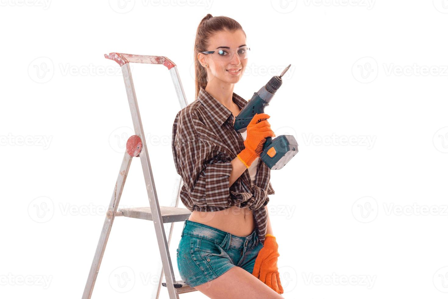 adorable young slim builder girl in glasses makes renovations with drill in her hands isolated on white background photo