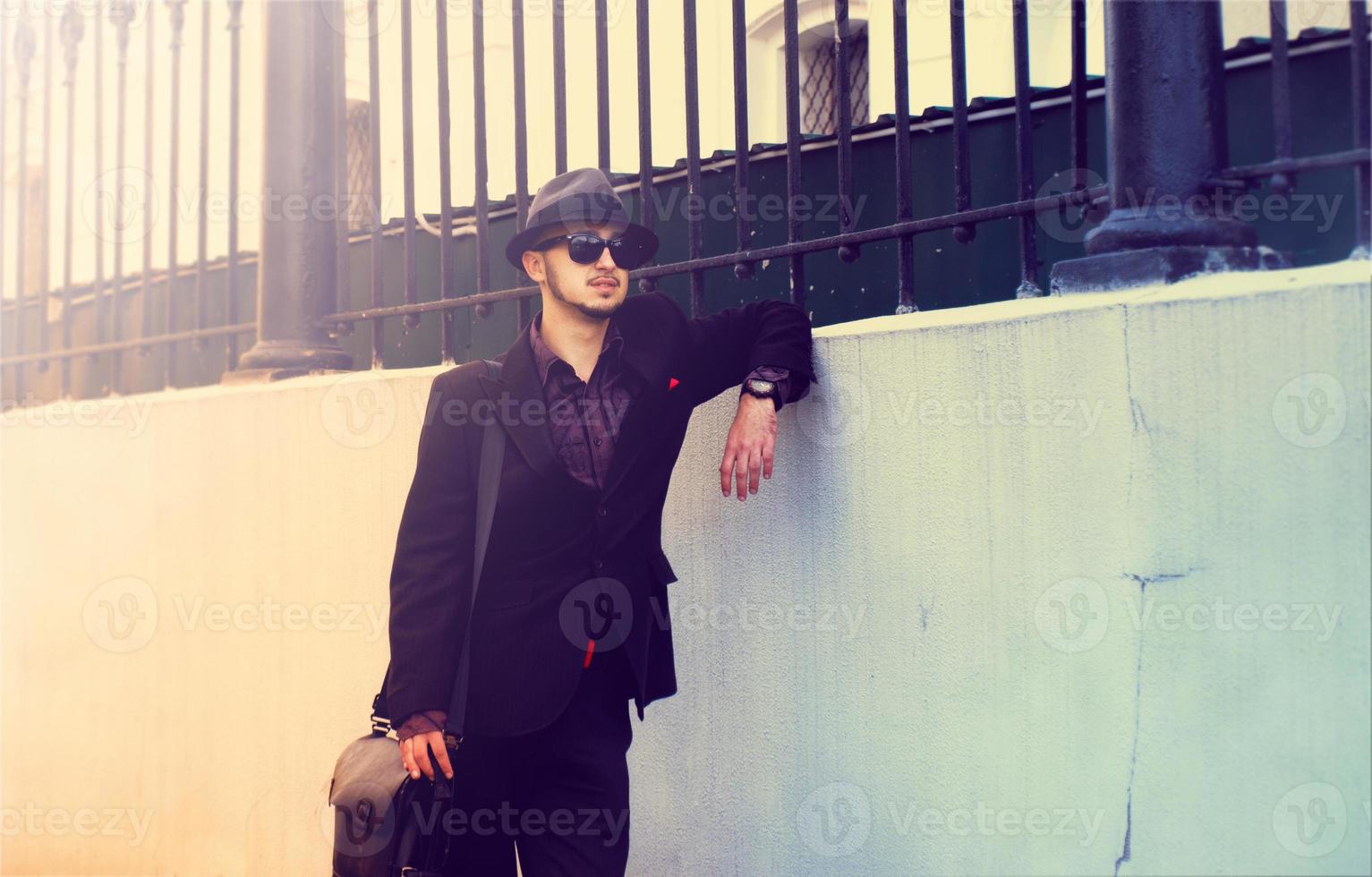 man in a hat, suit and glasses standing near the wall photo