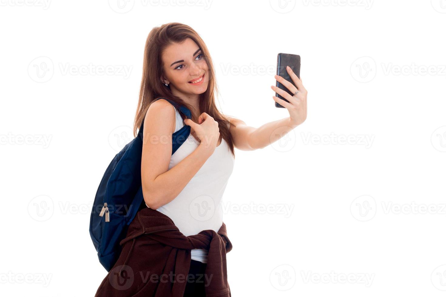 Una joven estudiante hermosa con mochila hace selfie aislada de fondo blanco en el estudio foto