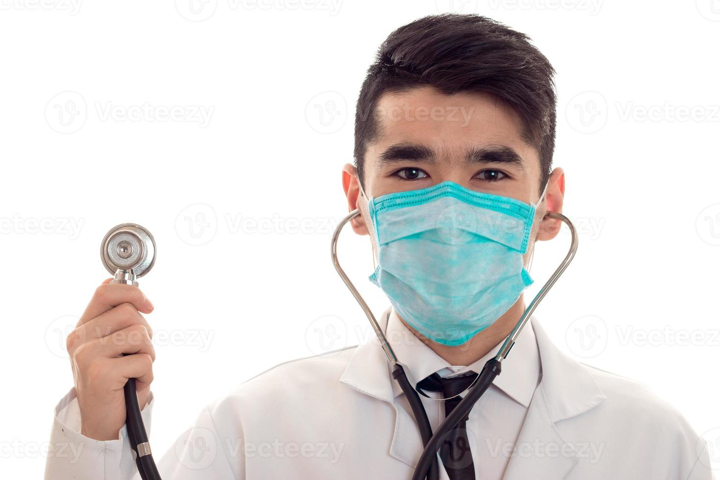 Joven médico apuesto posando en uniforme y máscara con estetoscopio aislado en fondo blanco en el estudio foto