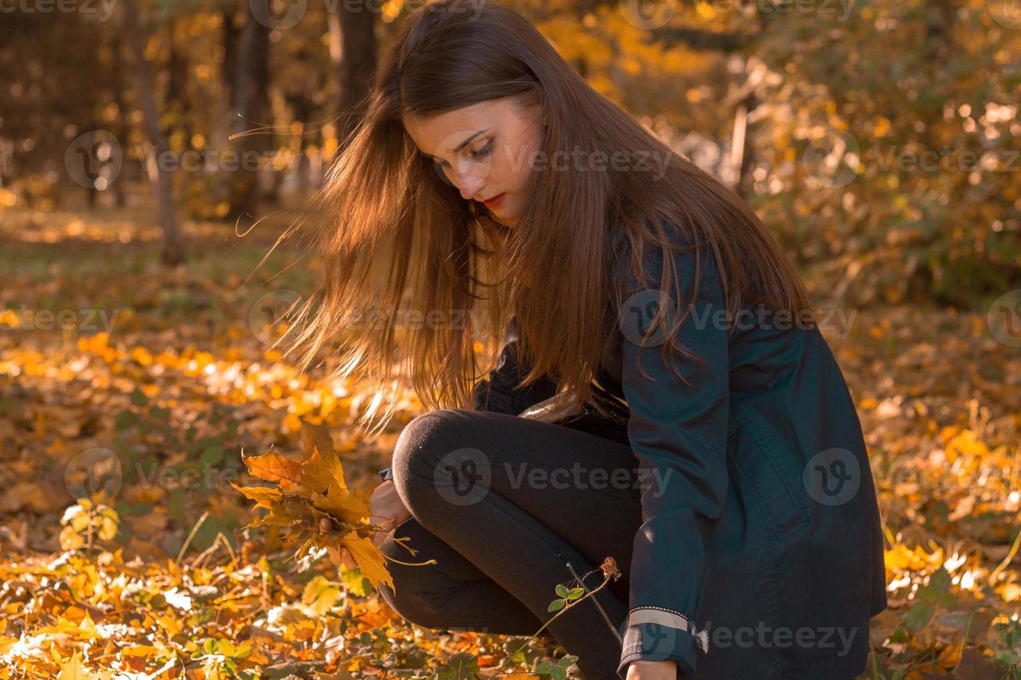 niña con el pelo largo sentada en el césped entre las hojas en el parque de otoño foto