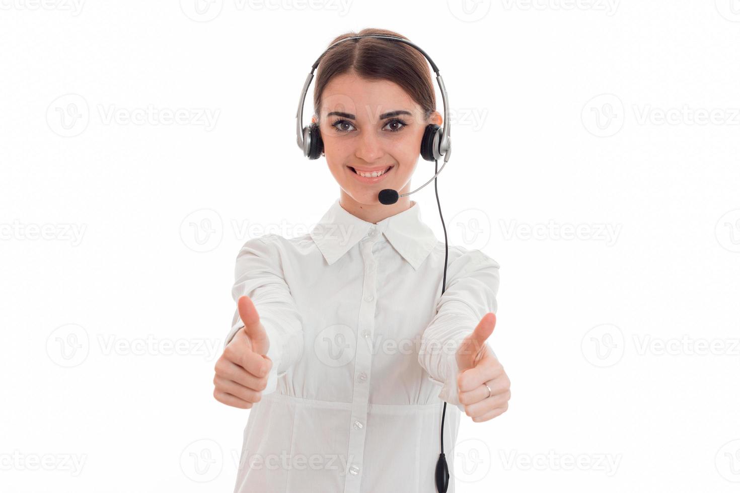 Bonita morena joven trabajadora de oficina de llamadas con auriculares y micrófono sonriendo y mostrando los pulgares hacia arriba en la cámara aislada en el fondo blanco foto