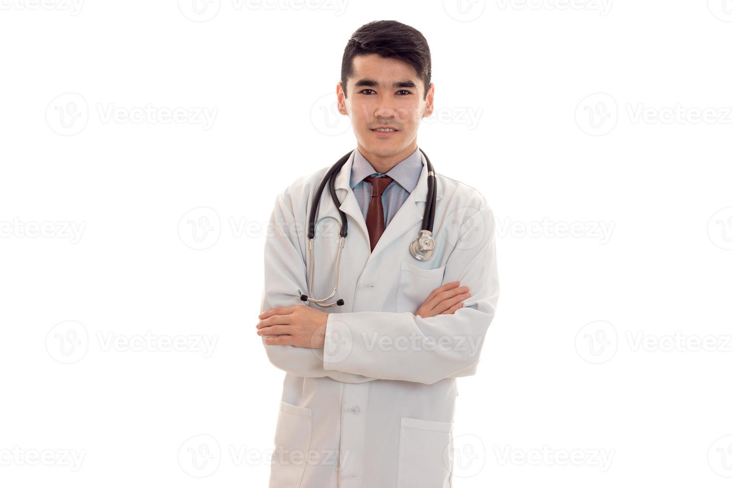 apuesto joven moreno doctor en uniforme blanco con estetoscopio mirando y sonriendo en cámara aislada en estudio foto