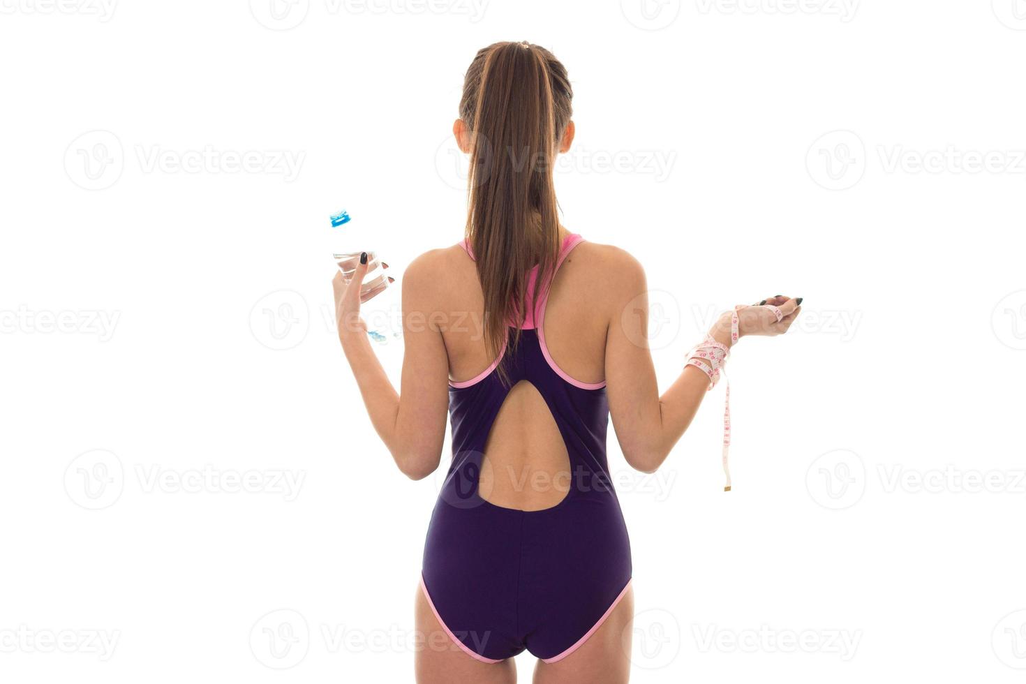 slim young brunette girl in body swimsuit with measure and bottle of water in hands from backside isolated on white background photo