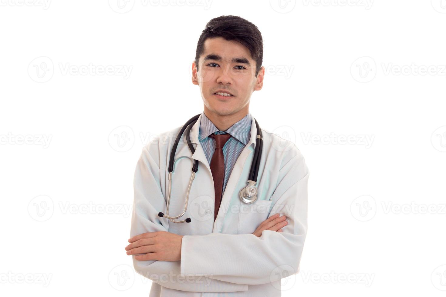 handsome young brunette man doctor in white uniform with stethoscope and crossed hands isolated in studio photo