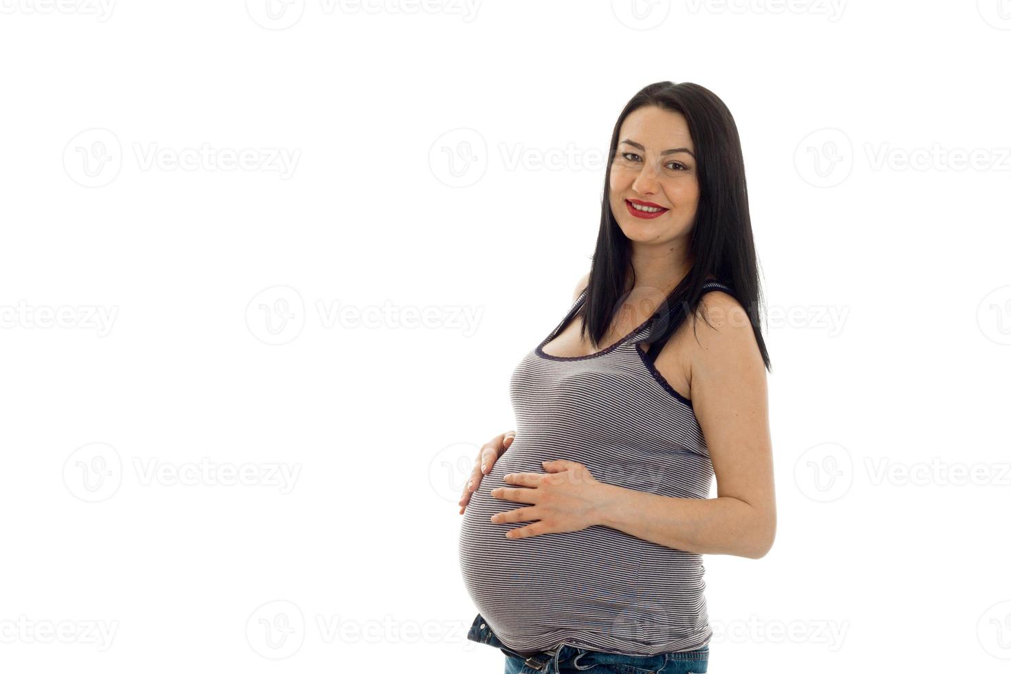 studio portrait of young pregnant brunette woman in shirt touching her belly and smiling on camera isolated on white background photo