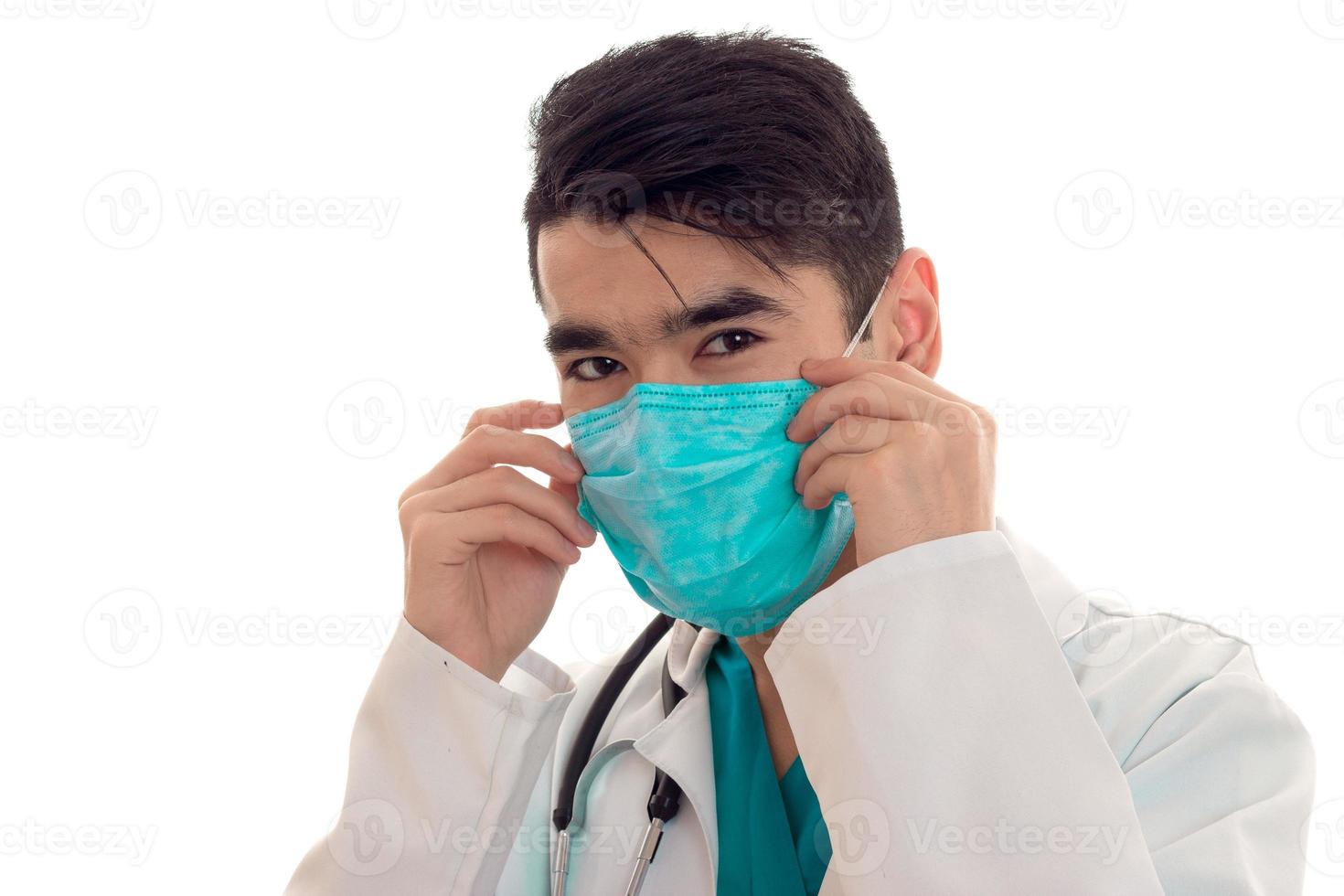 portrait of young handsome brunette man doctor in white uniform and blue mask with stethoscope looking at the camera isolated on empty background photo