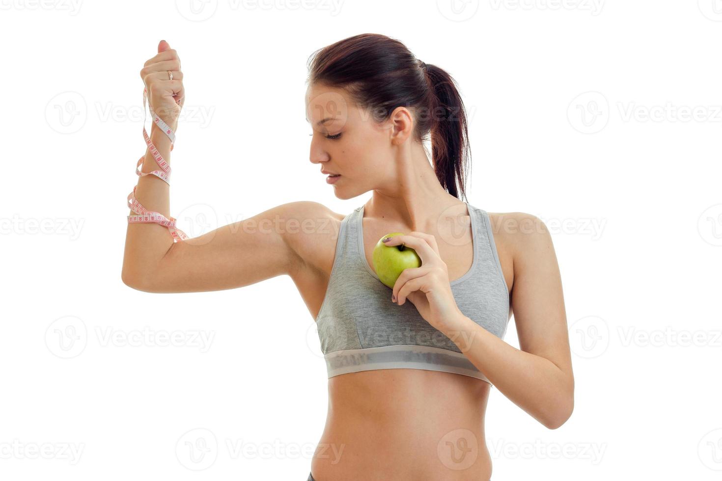 a young athletic girl in grey top with a tape measure on hand holds an Apple and looks toward photo