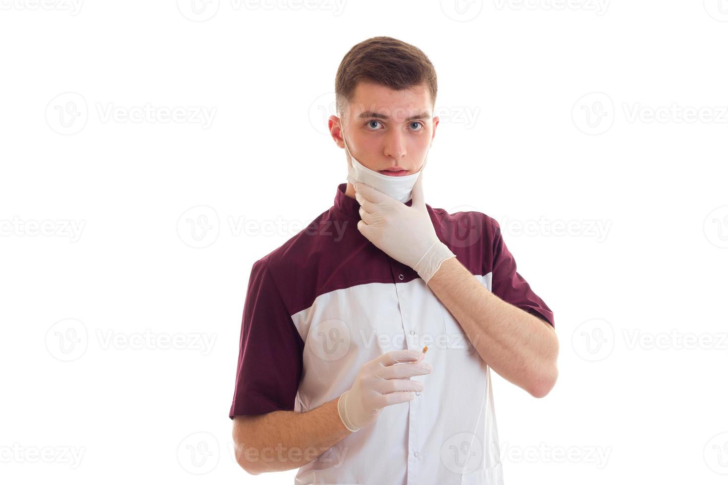 un joven médico serio sosteniendo soportes de jeringas con una máscara en la cara y mirando directamente foto