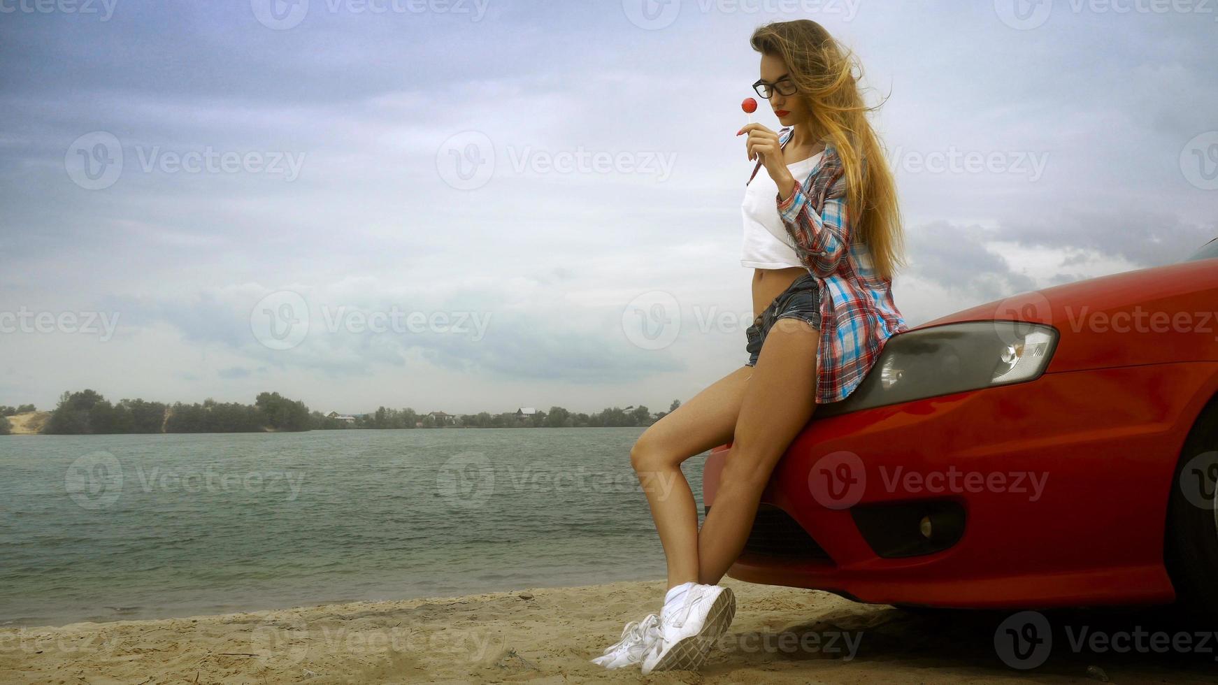 sensual blonde in glasses sits on a red car with lollipop in her hands photo