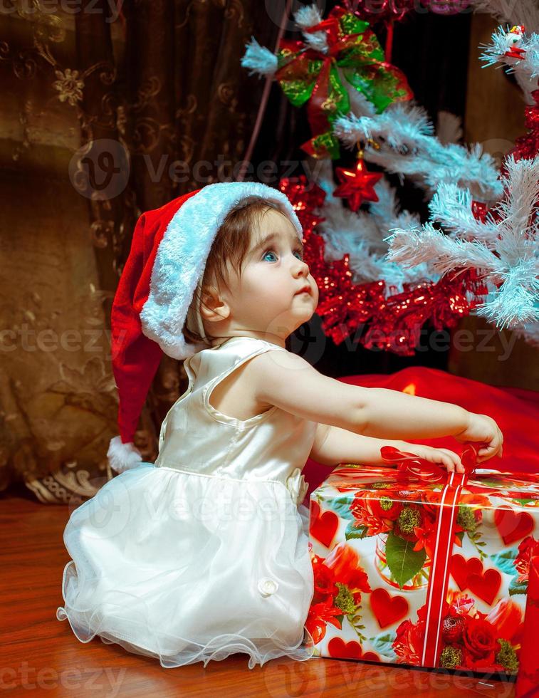 Cute little baby girl in Stana hat opens her first Christmas present photo