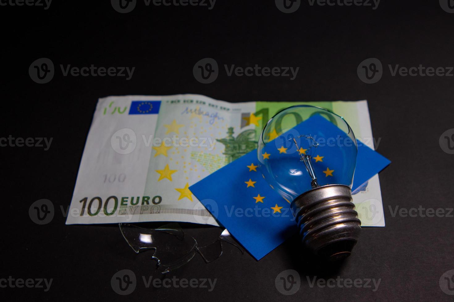 Broken light bulb with pieces of glass on 100 Euro bill and flag Europe. Concept of electricity prices and the electricity crisis in Europe photo