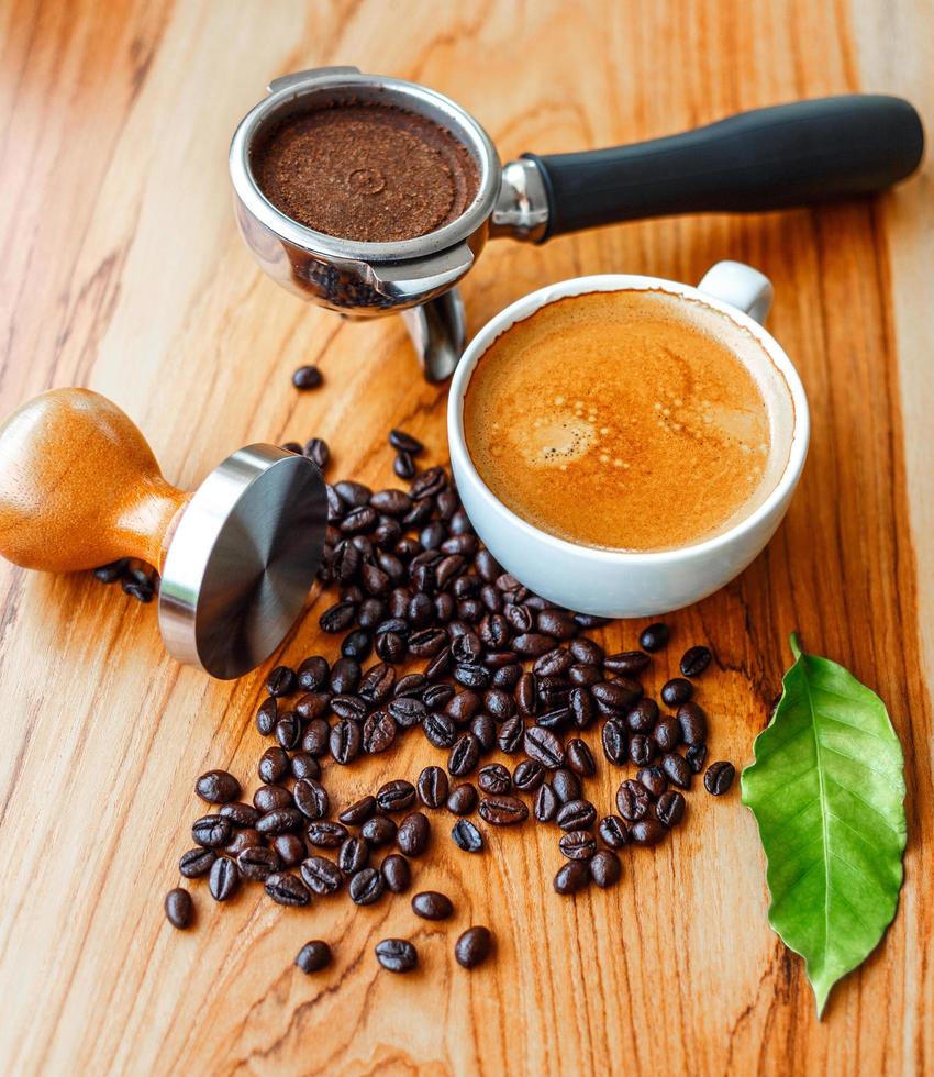 Top view of Espresso coffee cup and equipment of barista coffee tool portafilter with tamper and dark roasted coffee beans with green coffee leaf  on wooden table photo