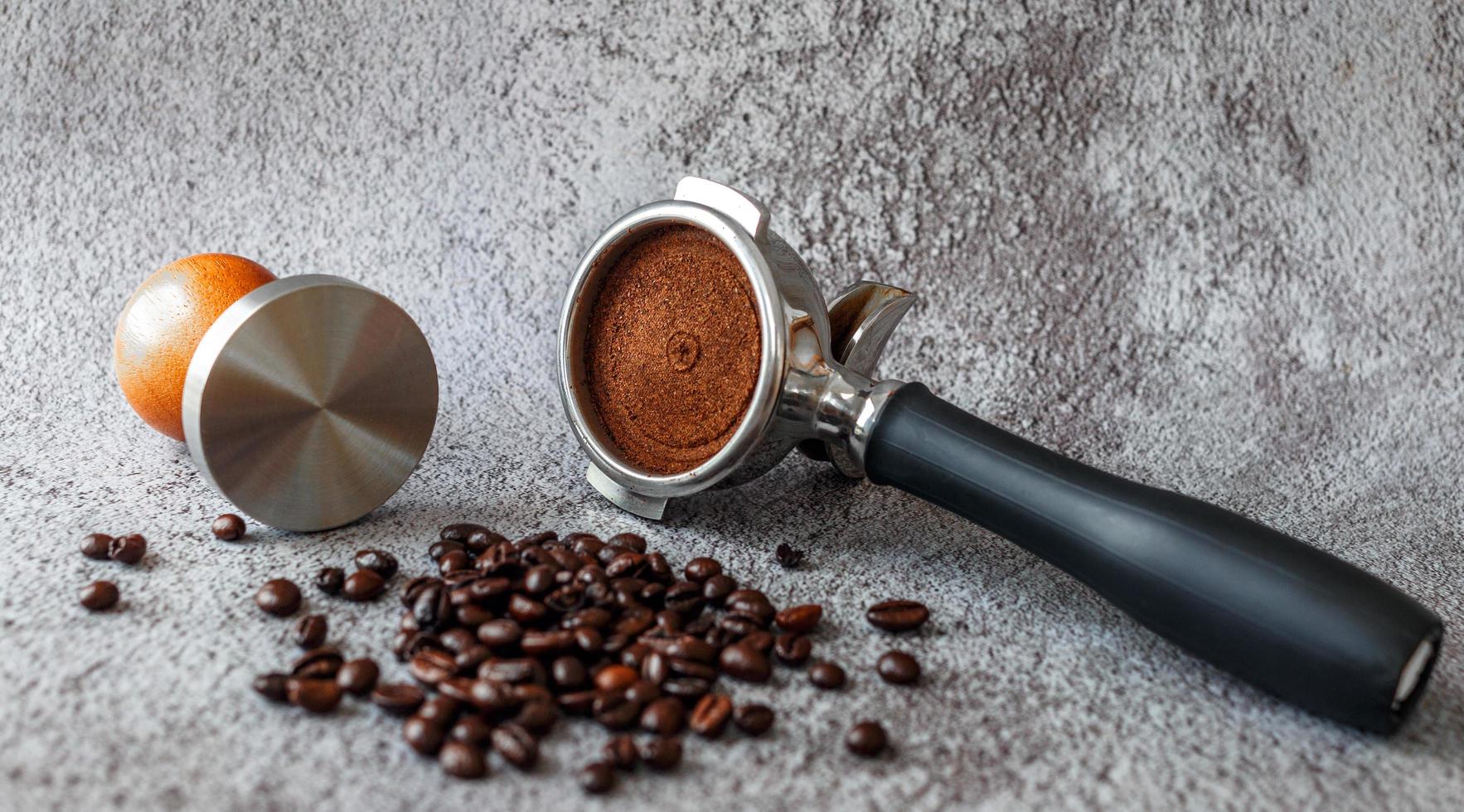 Equipment in a coffee shop of barista coffee tool portafilter with tamper and dark roasted coffee beans on gray background photo
