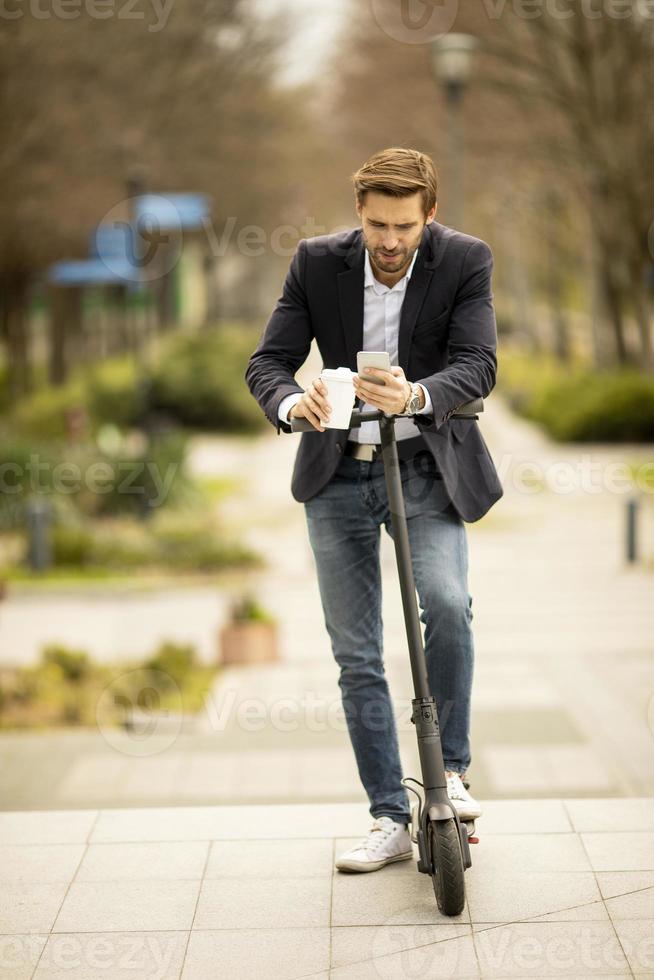 Young businessman using mobile phone while holding take away coffee cup on electric scooter photo