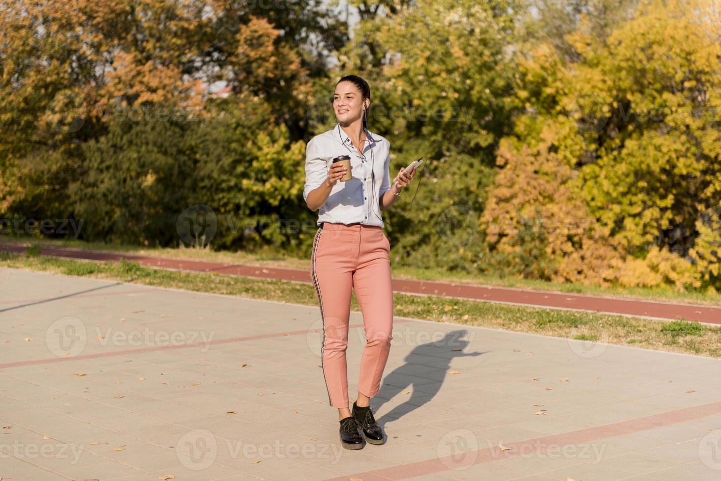 Happy young woman with coffee listening music on smartphone and walking on promenade photo