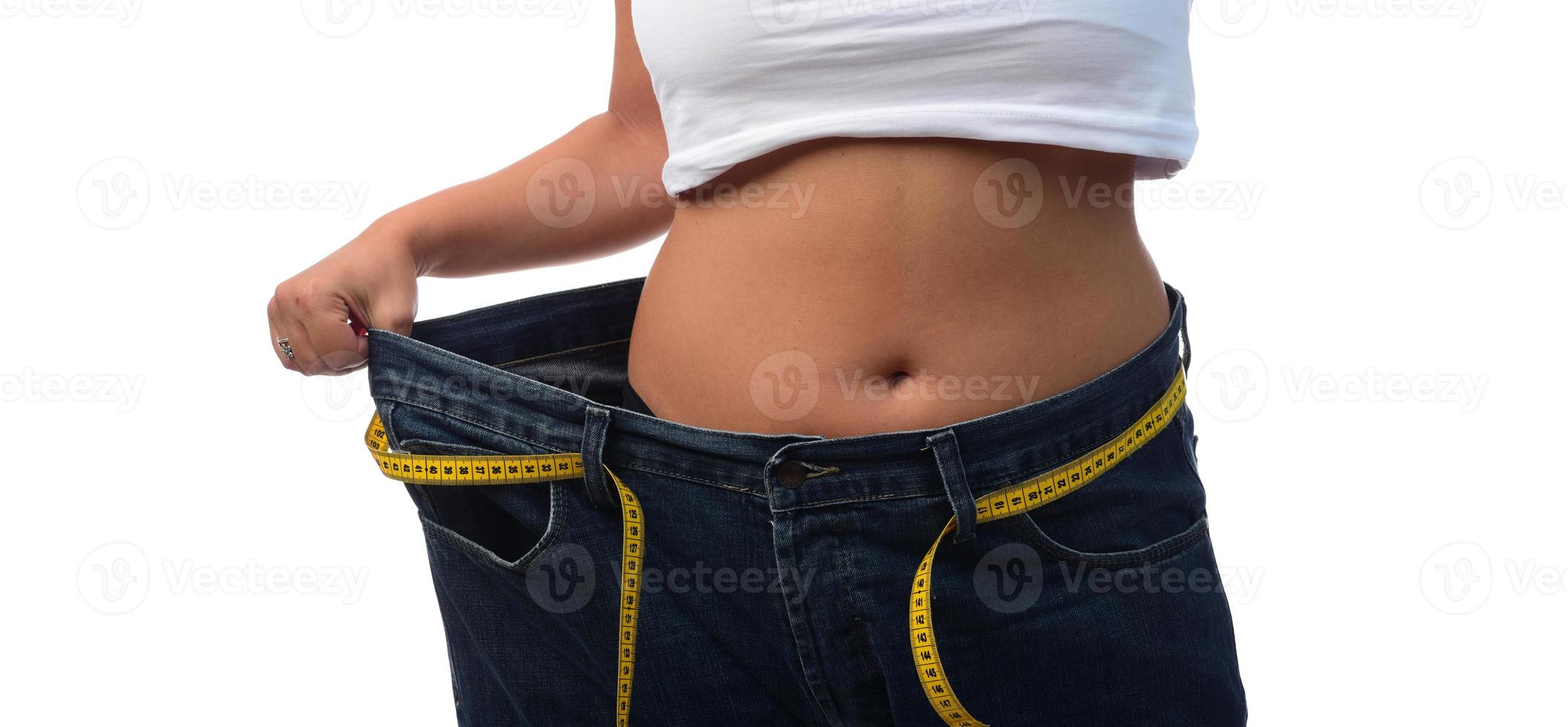 mid section of a woman with belly fat holding her blue jeans. Girl with big jeans isolated on white background. Woman shows her weight loss. Healthy lifestyle concept photo