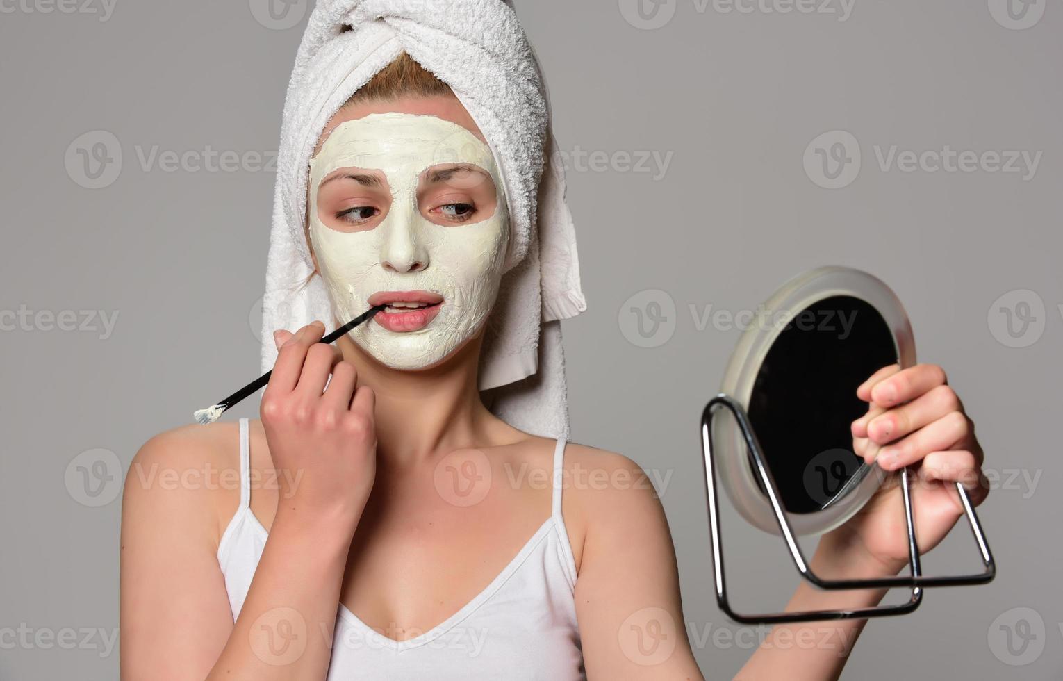 Beautiful female model with towel on her hear and white facial cosmetic mask on her face. Beauty cosmetic concept. Isolated on gray background. photo