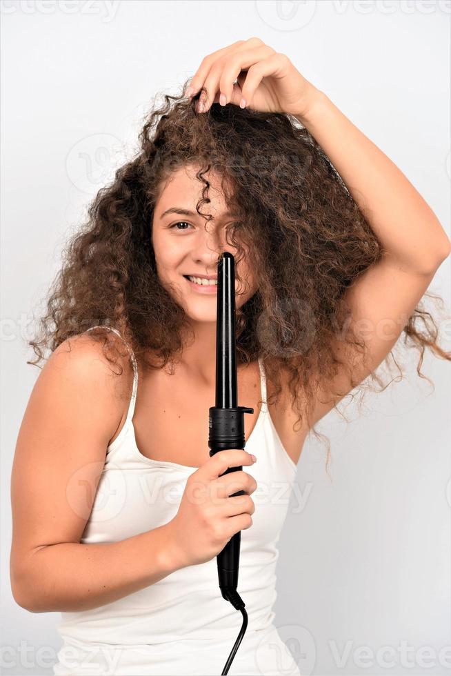 Curly Hair. Beautiful Smiling Woman With Long Wavy Hair Ironing It, Using Curling Iron. Happy Girl With Gorgeous Healthy Smooth Hair Using Curler For Perfect Curls. Hairstyle And Hairdressing photo