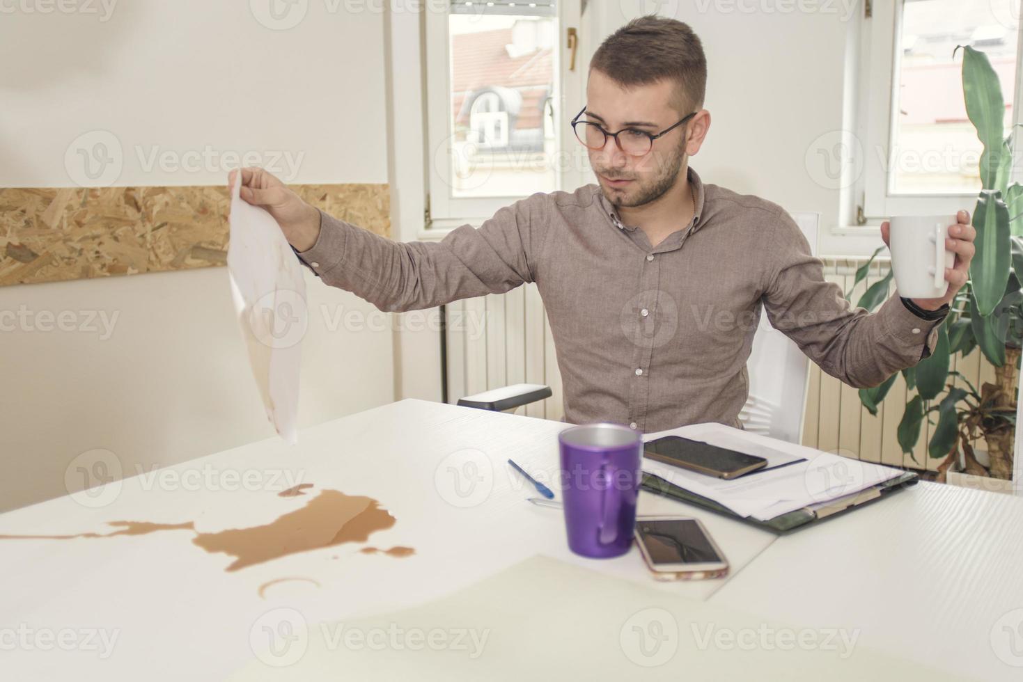 Scared and shocked young adult project manager in architect bureau working in professional design studio with blueprints, spilling coffee on documents, sitting behind table photo