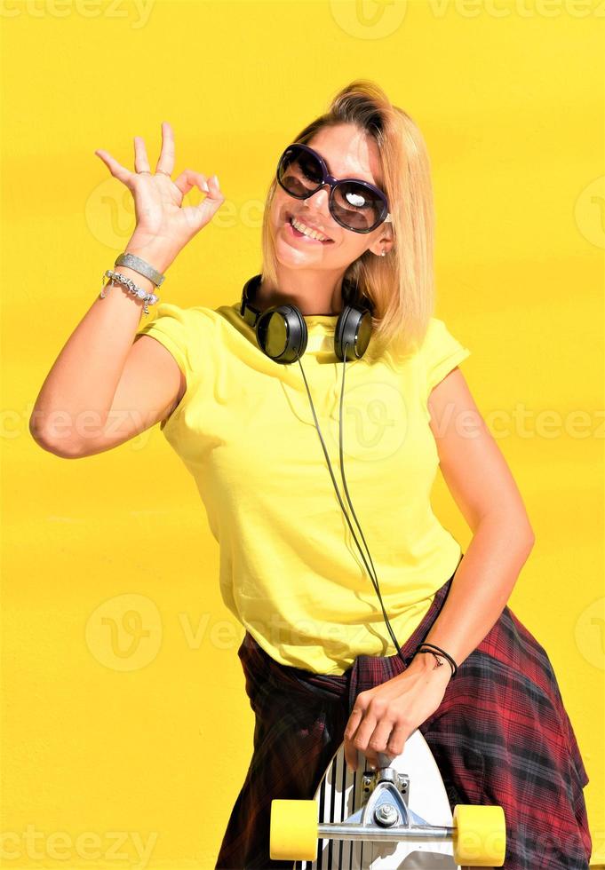 Portrait of happy beautiful blonde girl  hold her wooden longboard skateboard on yellow background wall, look to the camera and smile. Urban scene, city life. Hipster cute lady. photo