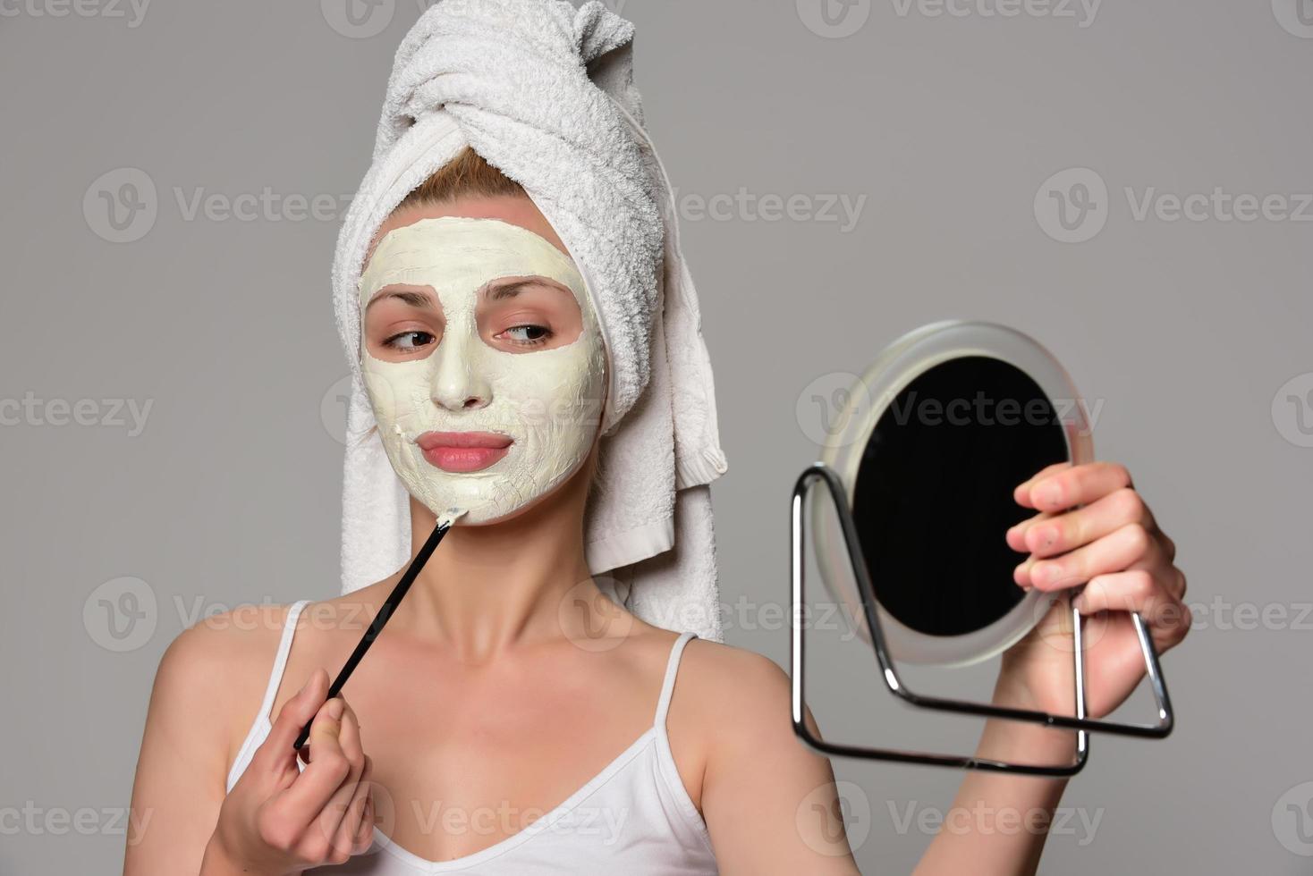 Beautiful female model with towel on her hear and white facial cosmetic mask on her face. Beauty cosmetic concept. Isolated on gray background. photo