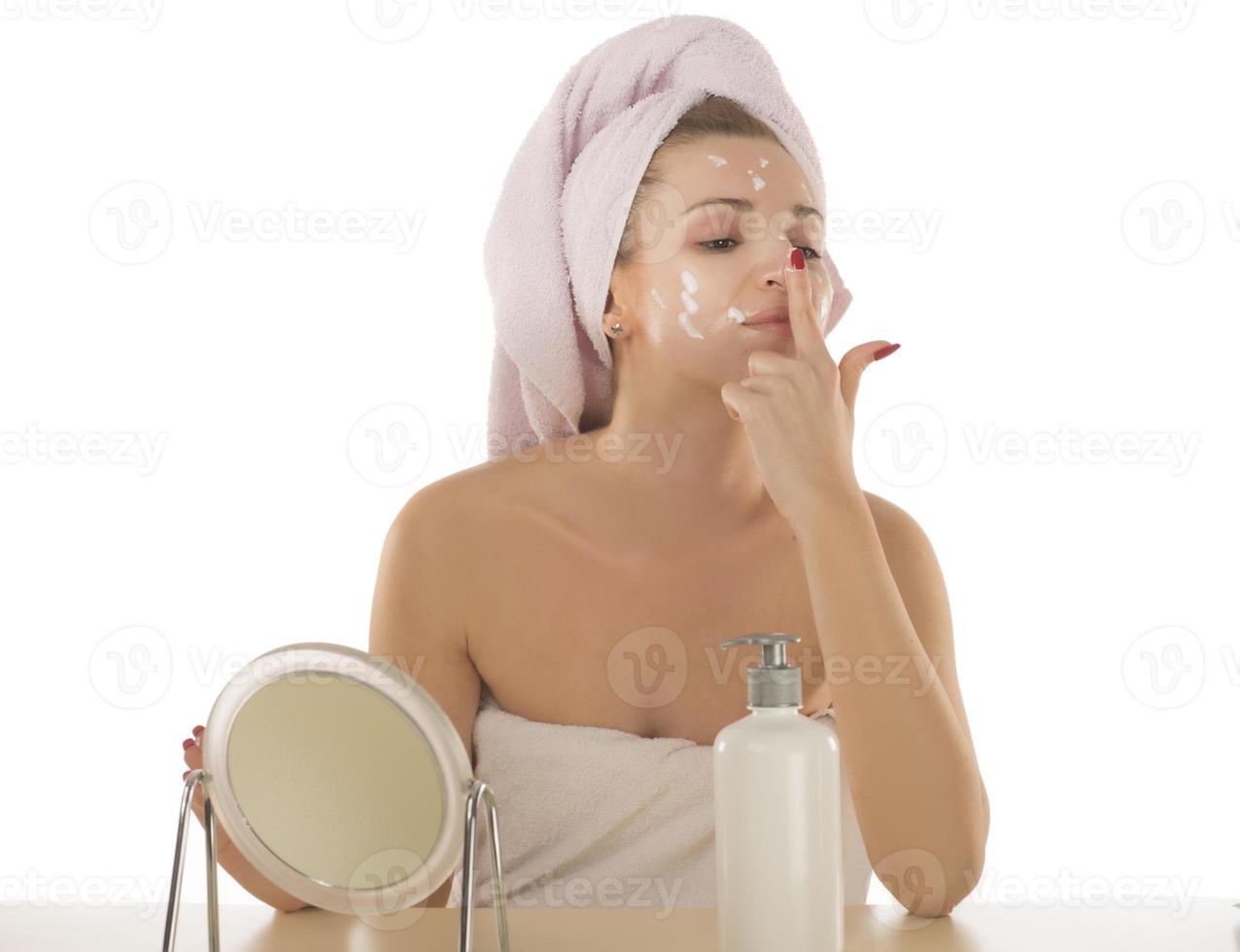 atractiva joven adulta aplica crema facial en el espejo, bella dama saludable envuelta en toallas pone crema hidratante de día nutritiva en la piel suave e hidratada en el baño foto