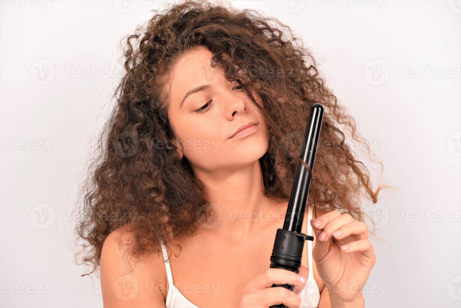 Beautiful Smiling Woman With Long Wavy Hair Ironing It, Using Curling Iron. Happy Girl With Gorgeous Healthy Smooth Hair Using Curler For Perfect Curls. Hairstyle And Hairdressing photo