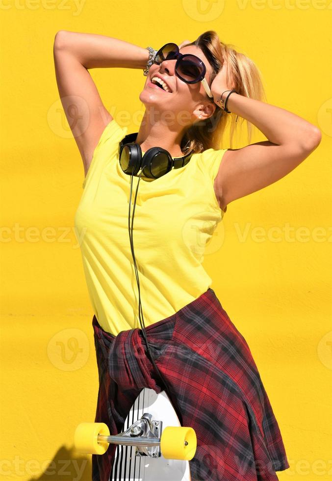 Portrait of happy beautiful blonde girl  hold her wooden longboard skateboard on yellow background wall, look to the camera and smile. Urban scene, city life. Hipster cute lady. photo