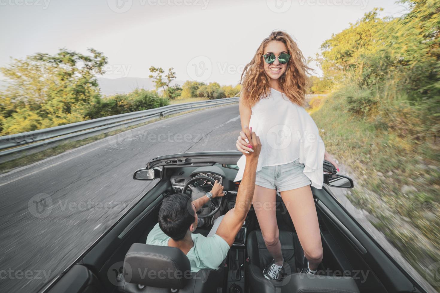 Friends having fun at car trip around the world. Couple in love with arms up on a convertible car. photo