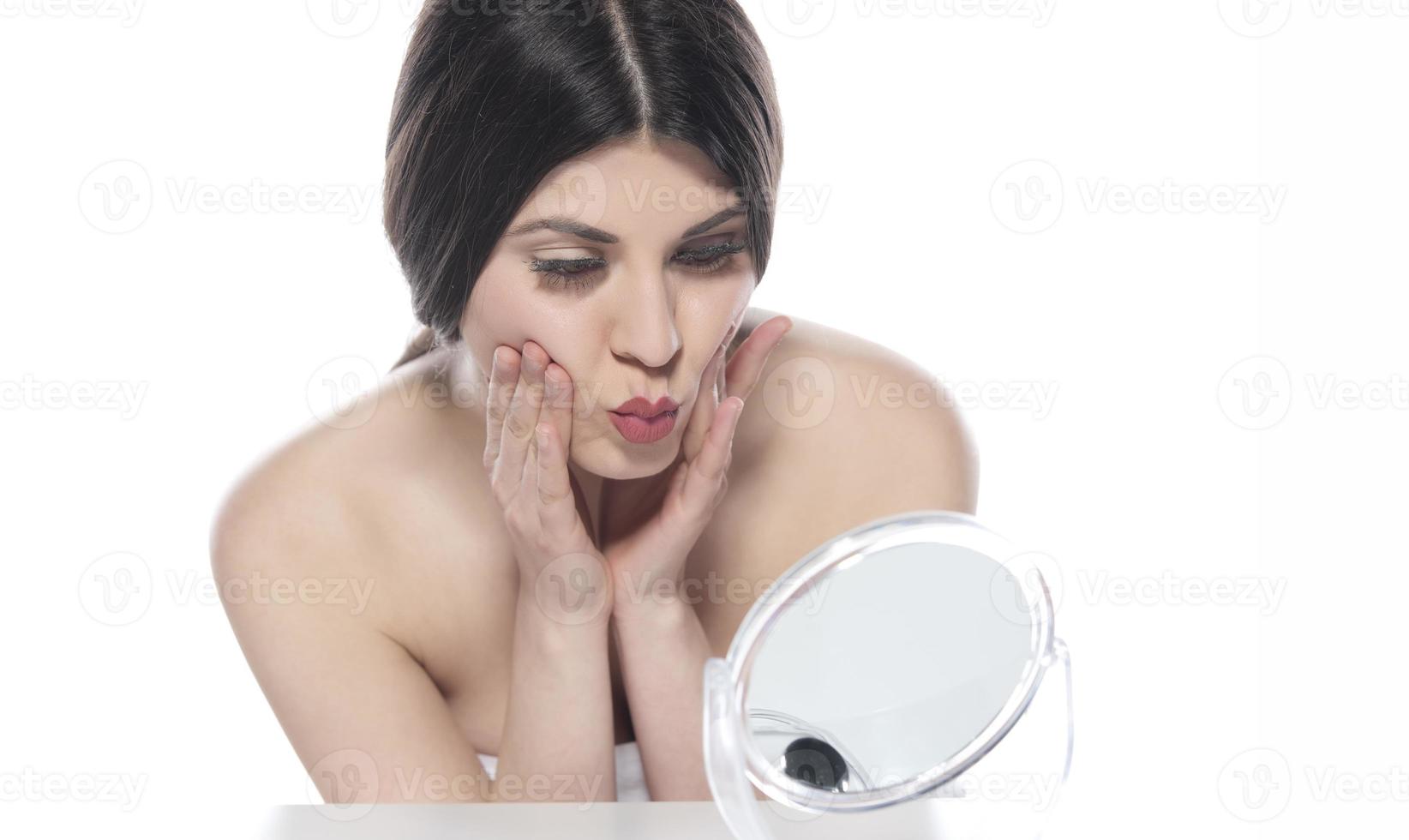 beautiful attractive brunette woman checking her face skin in front of a mirror on white background. photo