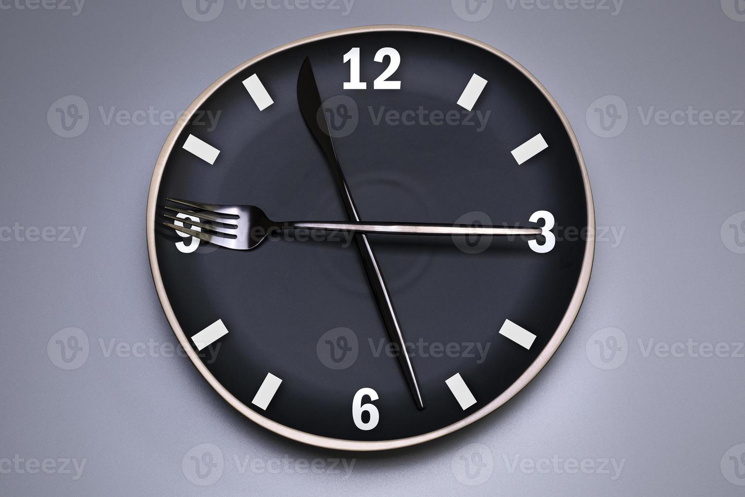 Clock on the black plate, with cutlery, grey background. The concept of intermittent fasting, lunchtime, diet and weight loss. photo